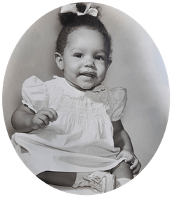 Young child in a white dress, sitting and smiling, with a bow in their hair, in a studio setting.