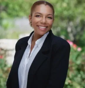 Woman in black blazer and white shirt smiles outdoors.