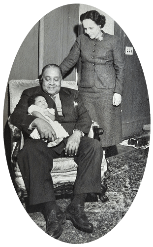 Man in suit holding a baby, woman in suit standing behind, indoor setting.