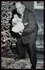 Man holding a baby, smiling. They stand near a Christmas tree and patterned rug.