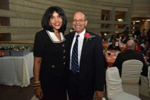 Woman in black dress and man in suit pose at an event, smiling. Tables and guests in background.