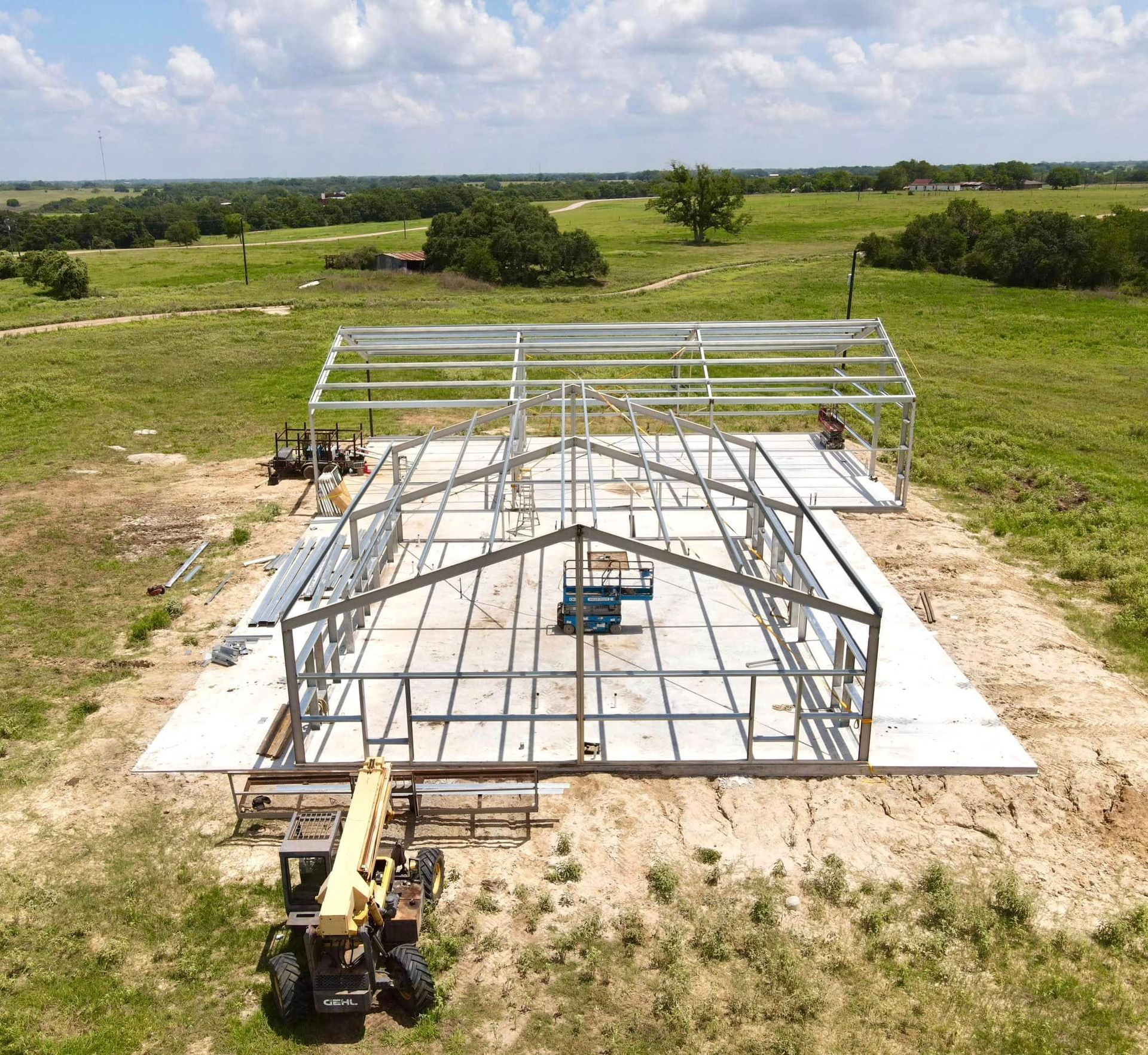 Steel frame of a building under construction on a concrete slab in a grassy field.