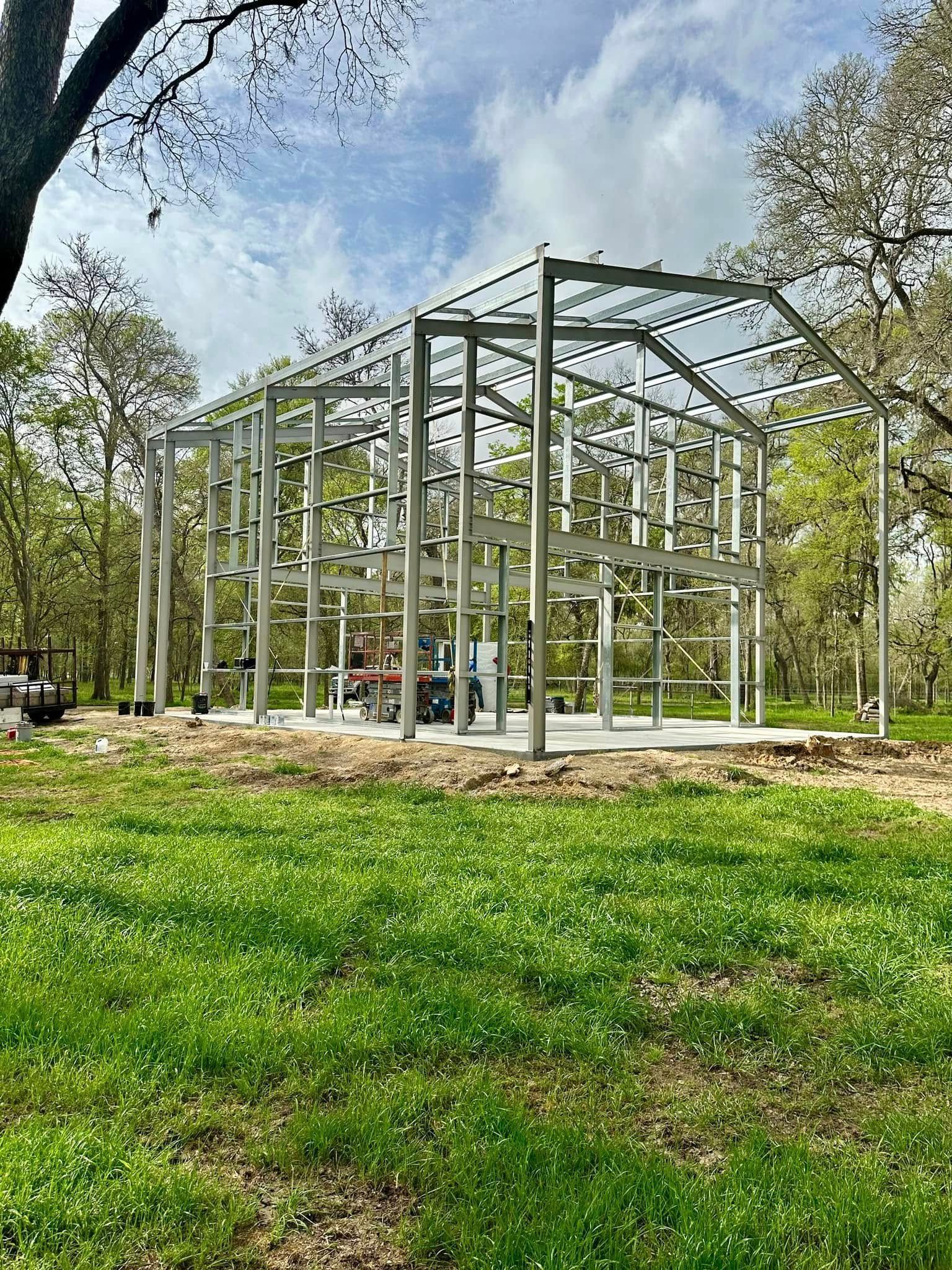 Metal framework of a building under construction, set in a grassy field with trees.