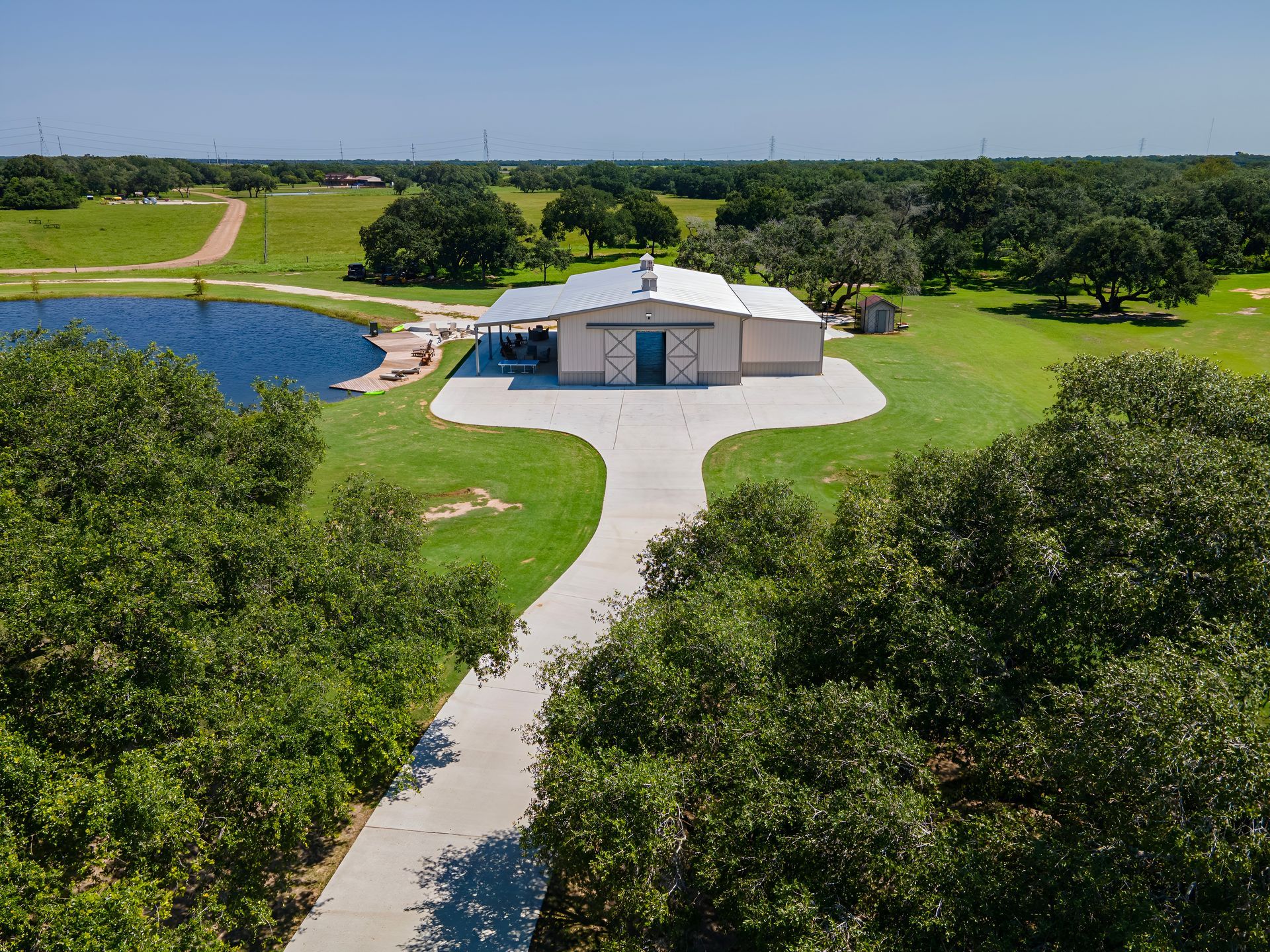 A barn-style building with a long driveway, pond, and green landscape on a sunny day.