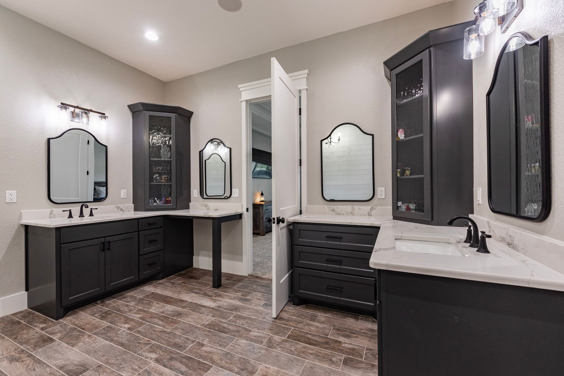 Dark gray bathroom with mirrors, cabinets, and white countertops. Doorway leads to another room.