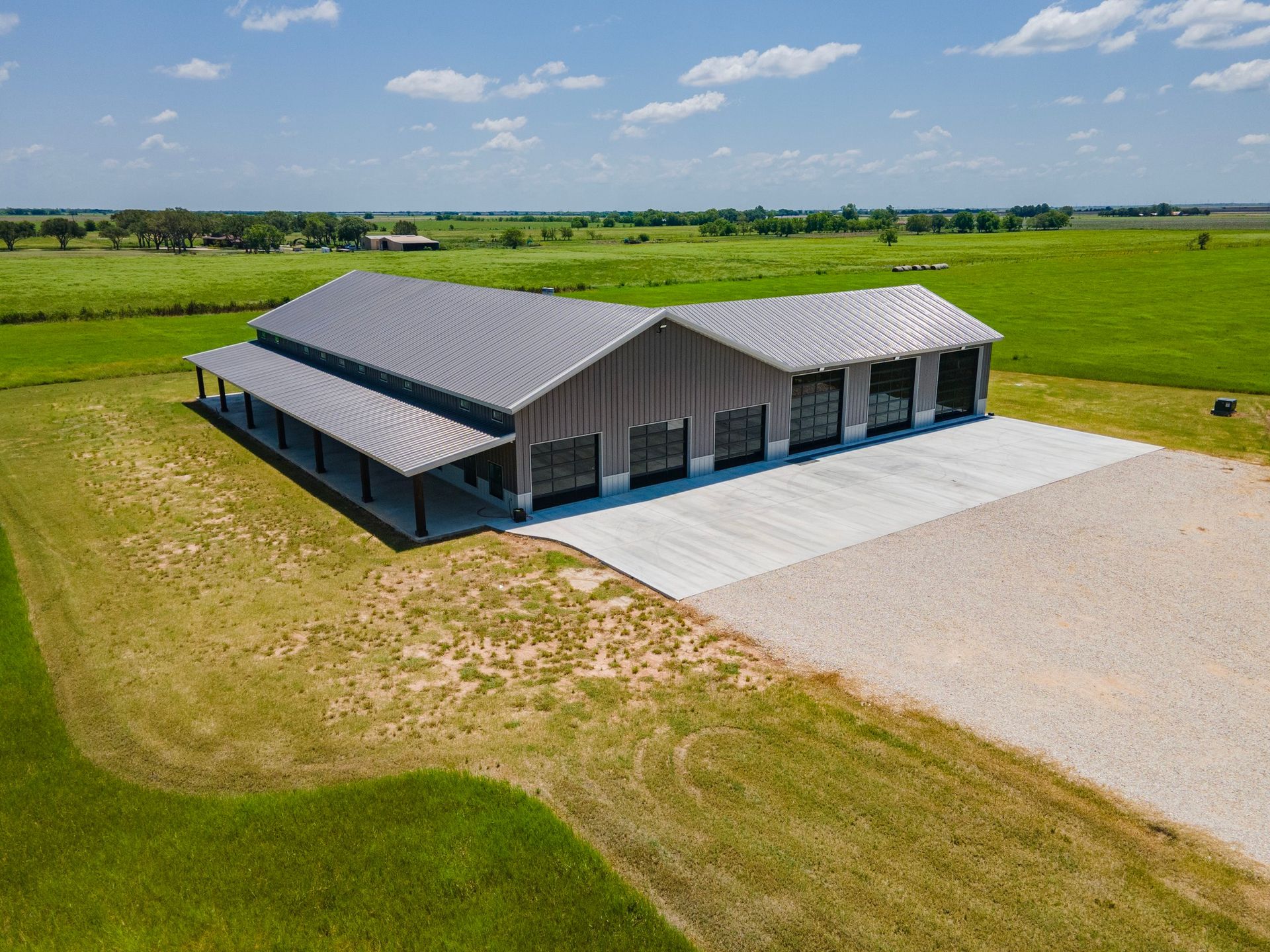 Large gray metal building with open-air sides on a concrete slab, surrounded by green fields and gravel.