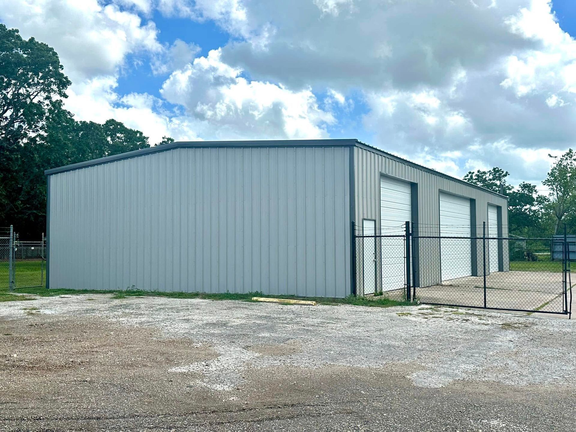 Gray metal building with three garage doors on a gravel lot under a cloudy sky.