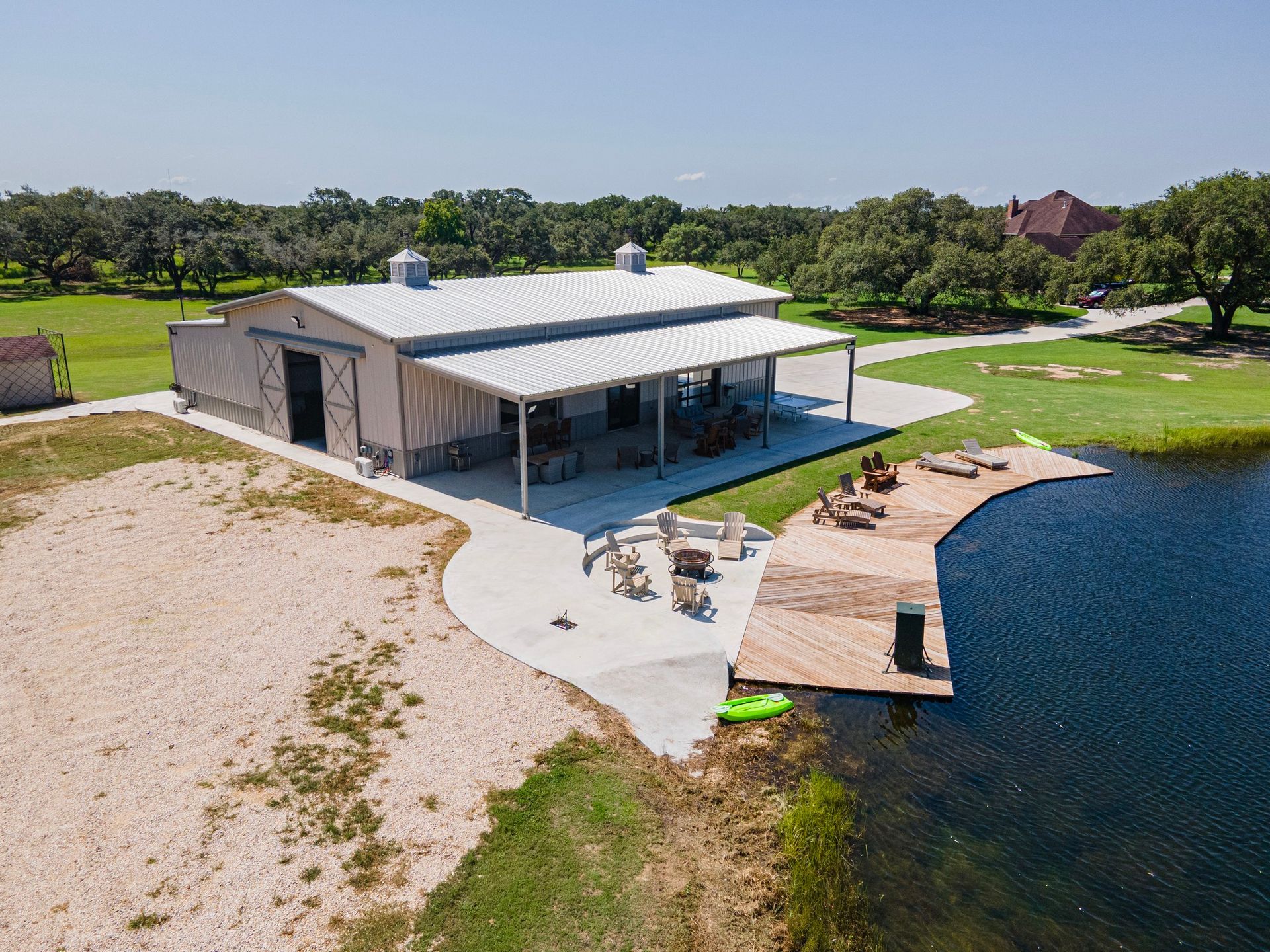 Barn-style building with porch and dock on a lake. Beige building with silver roof, lawn, trees, and blue water.