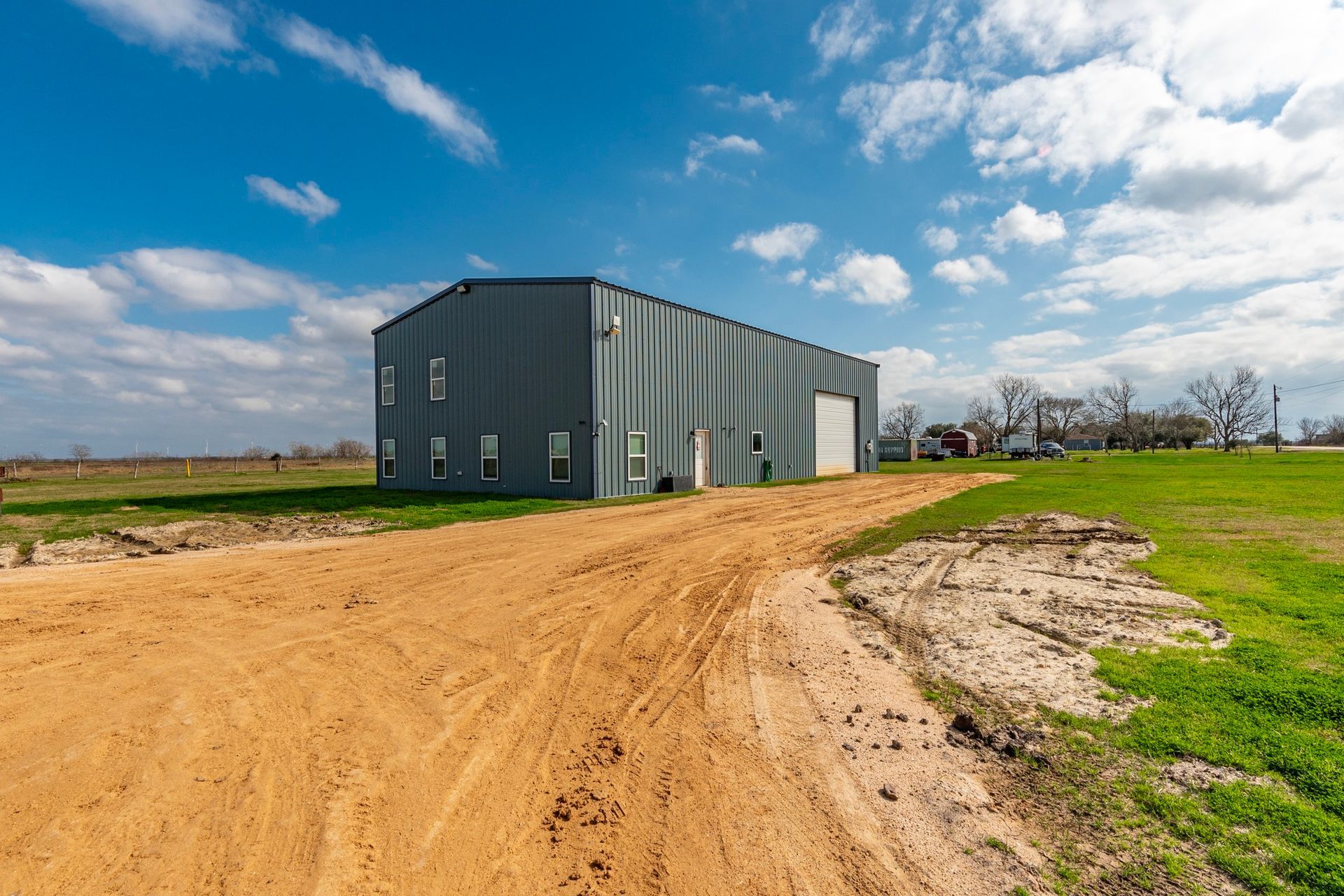 A gray industrial building on a sandy dirt road under a blue sky.