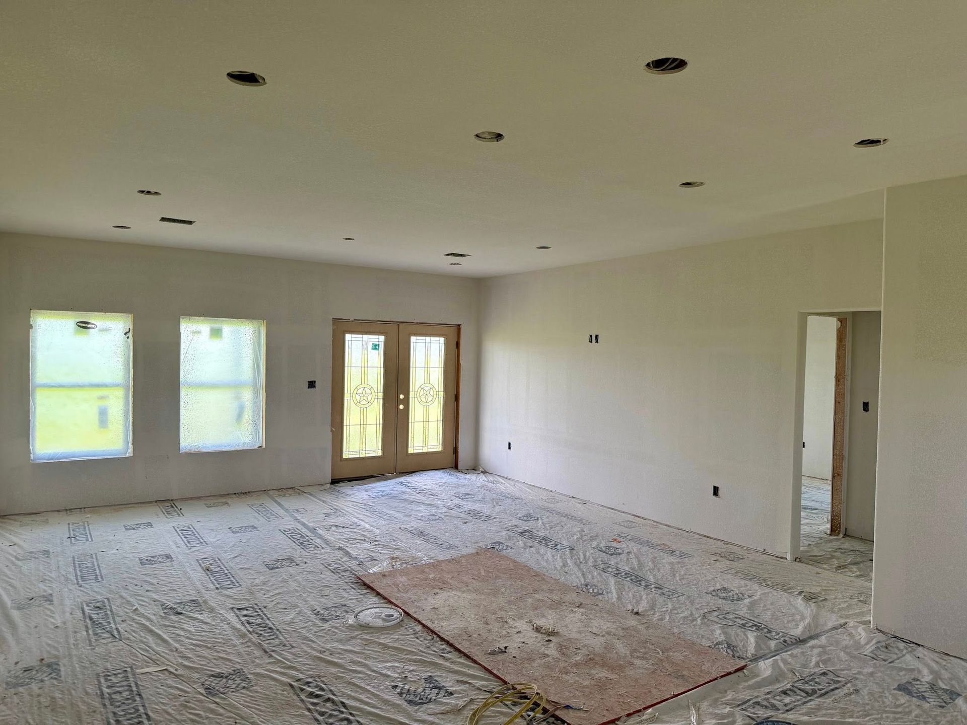 Interior room under construction with windows, doors, and recessed lighting. Drywall walls, plastic floor covering.