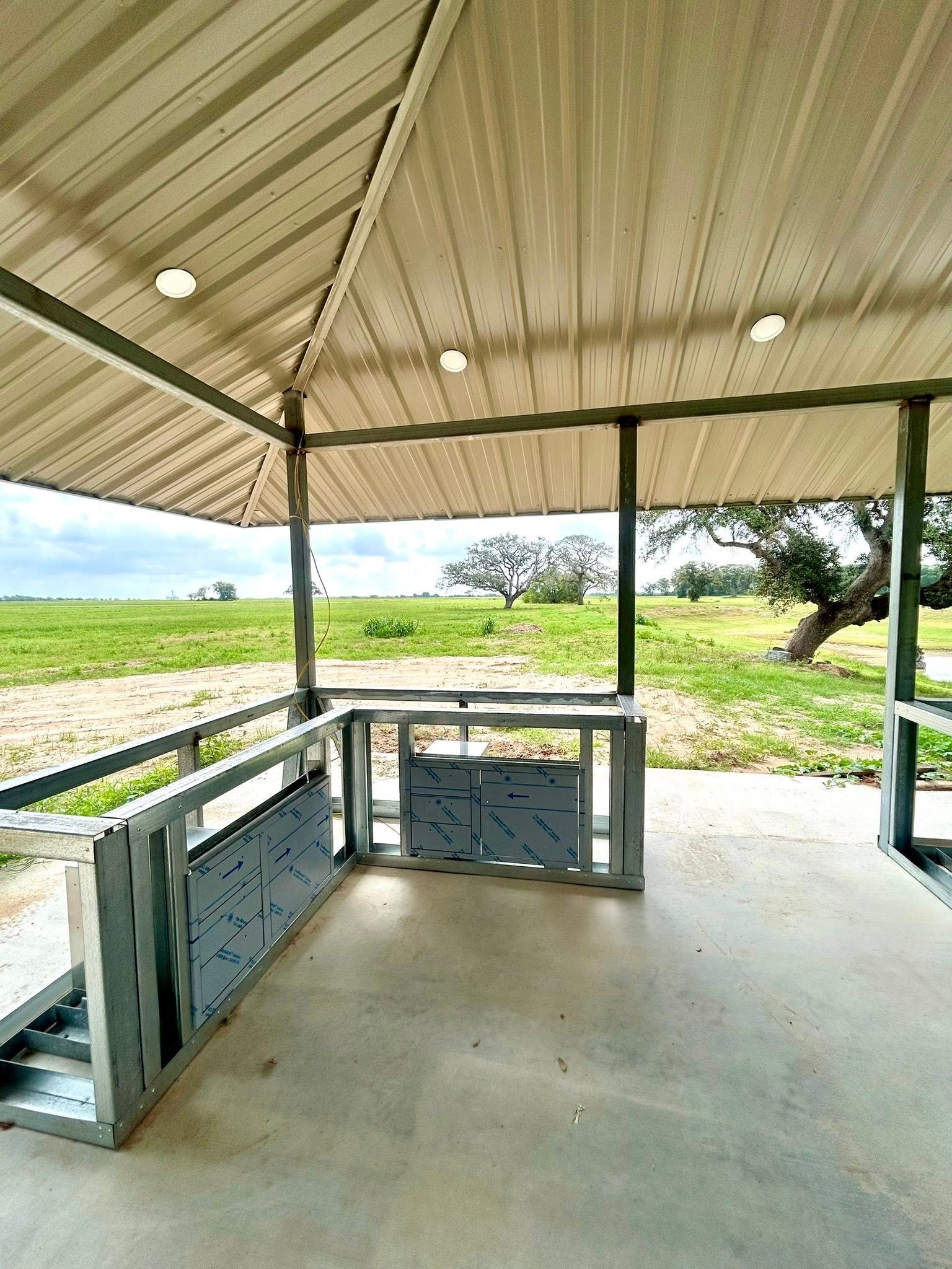 A metal framed outdoor pavilion with built-in grilling area overlooking a field and trees.