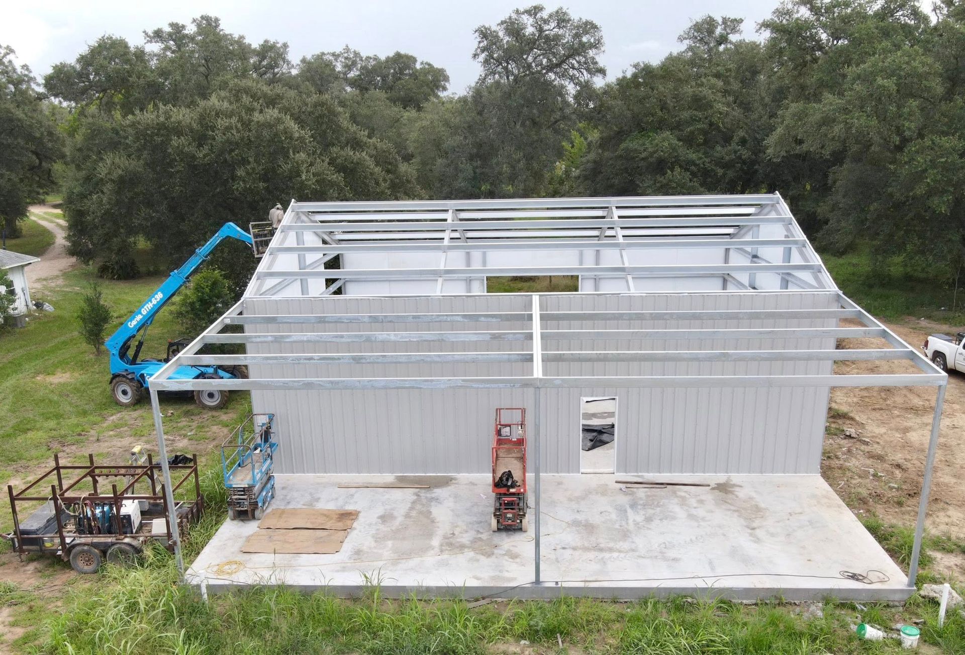 Construction of a metal building with a blue lift in a wooded area.