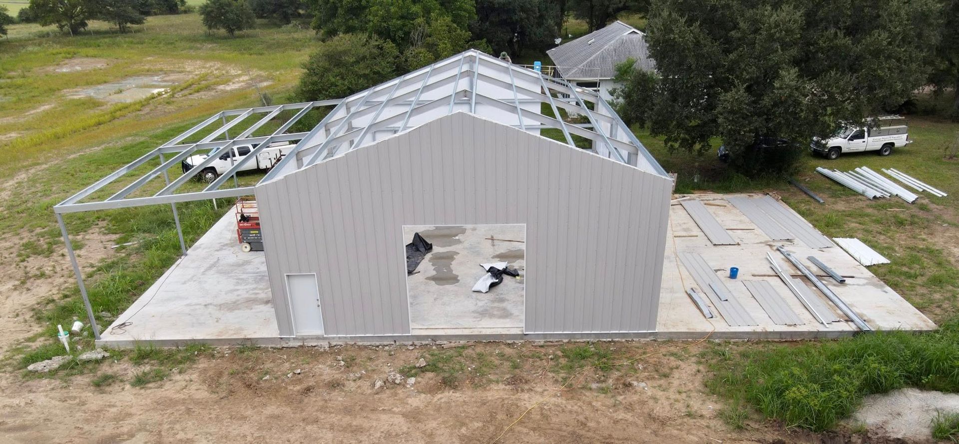 Construction of a gray metal building; framing, wall panels, and concrete foundation are visible. A vehicle is parked near the construction site.