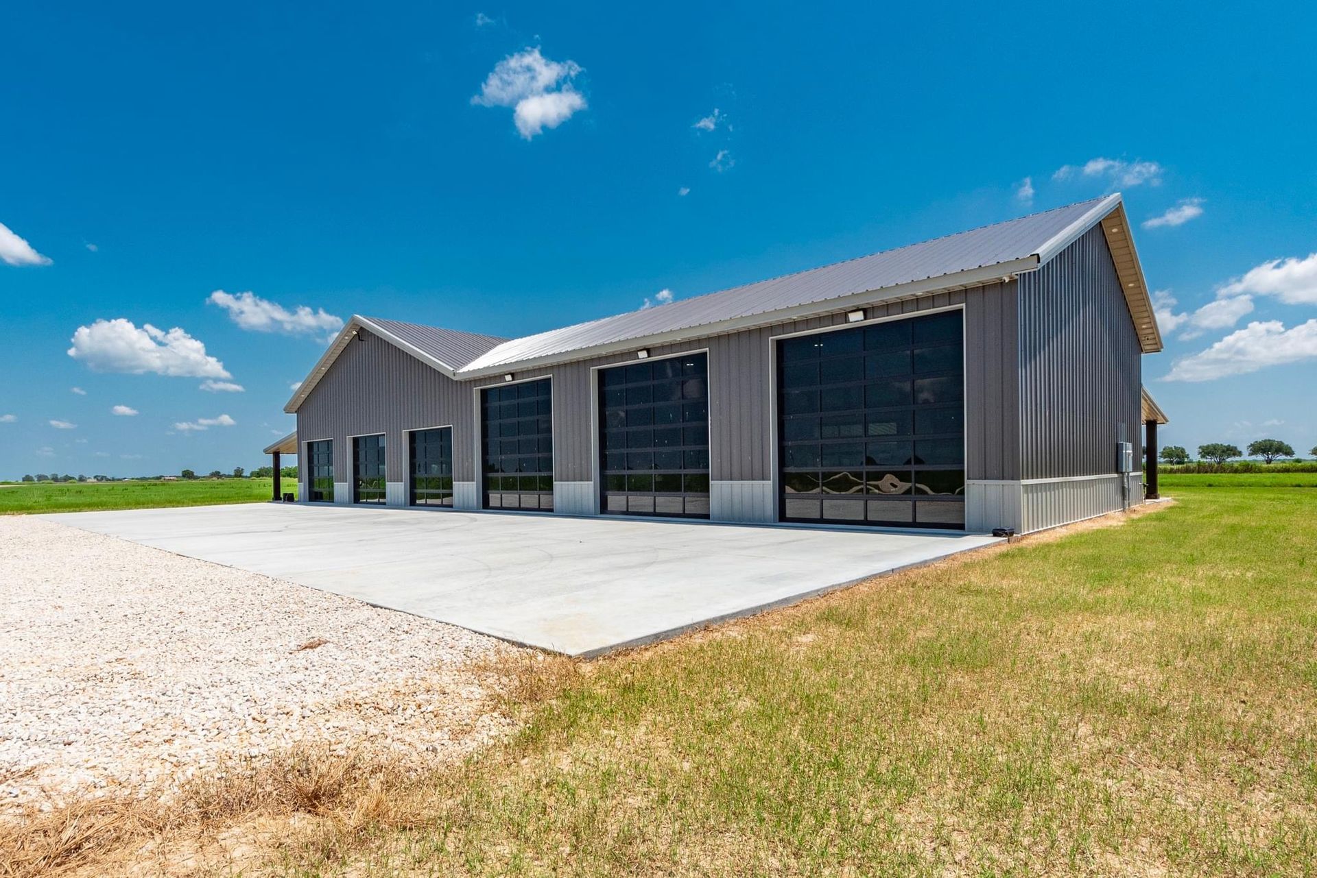 Gray metal building with large black garage doors on a concrete pad under a blue sky.