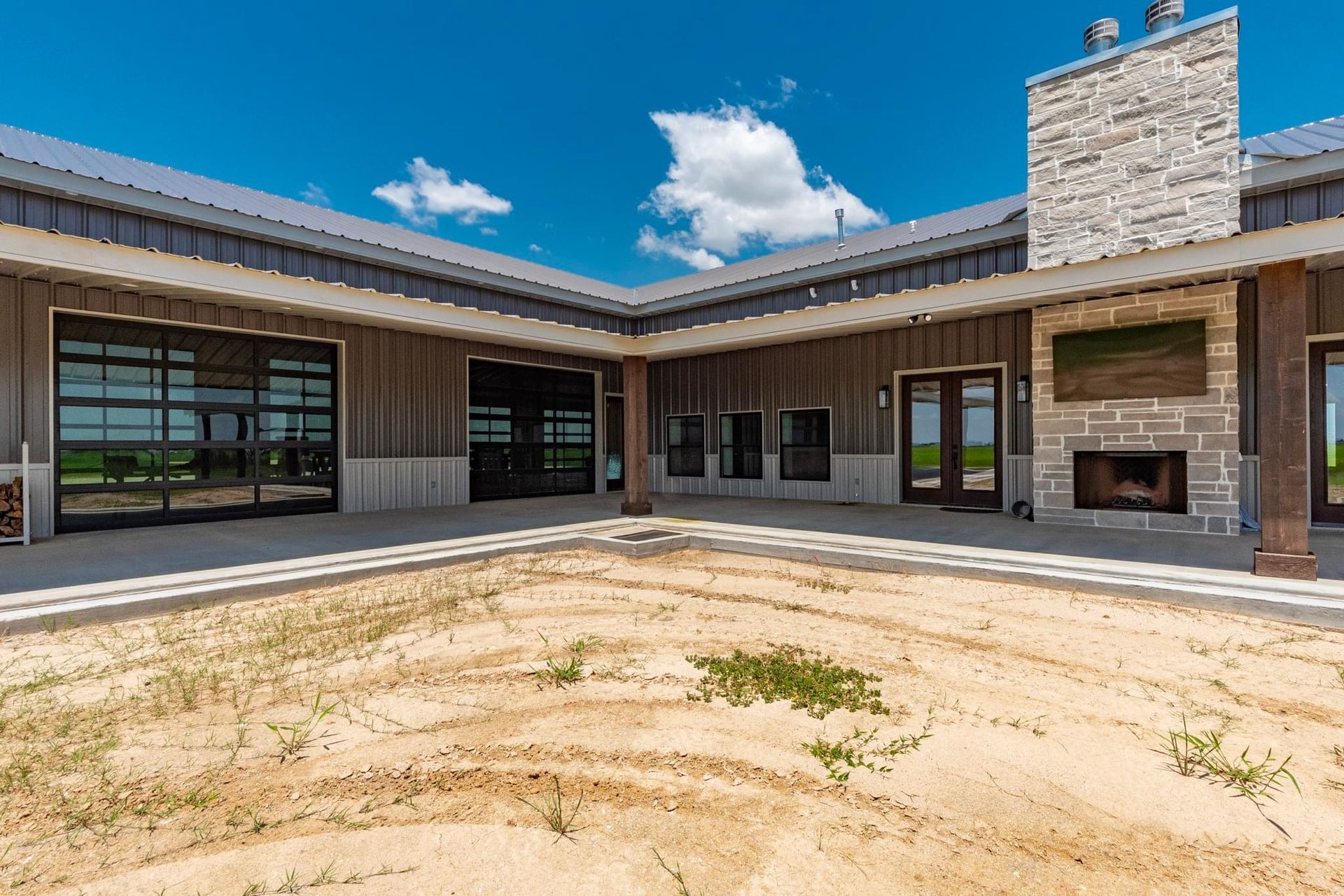 Exterior view of a modern building with glass garage doors, a stone fireplace, and a courtyard on a sunny day.