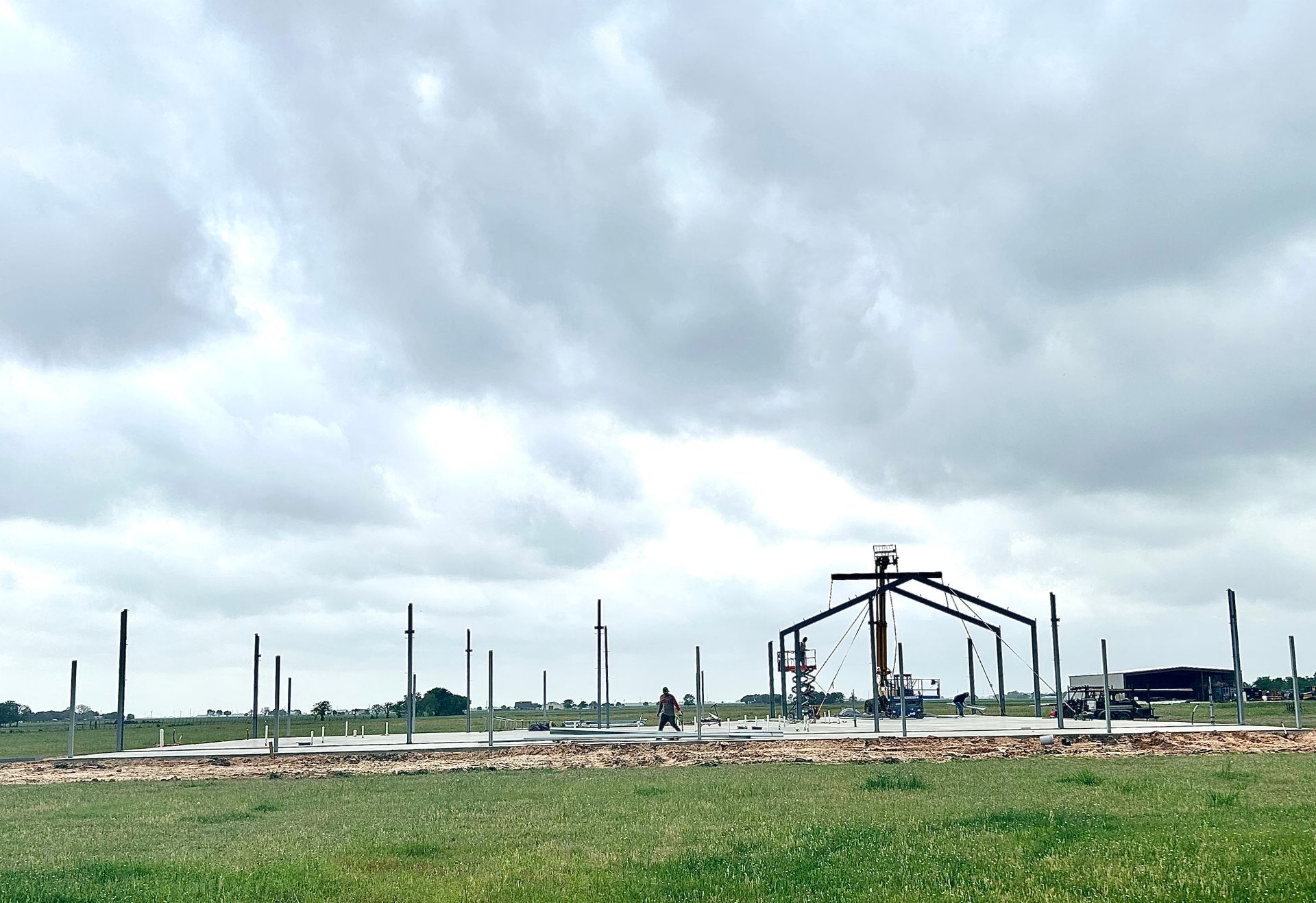 Construction site under cloudy sky with steel structures and concrete foundation in a field.