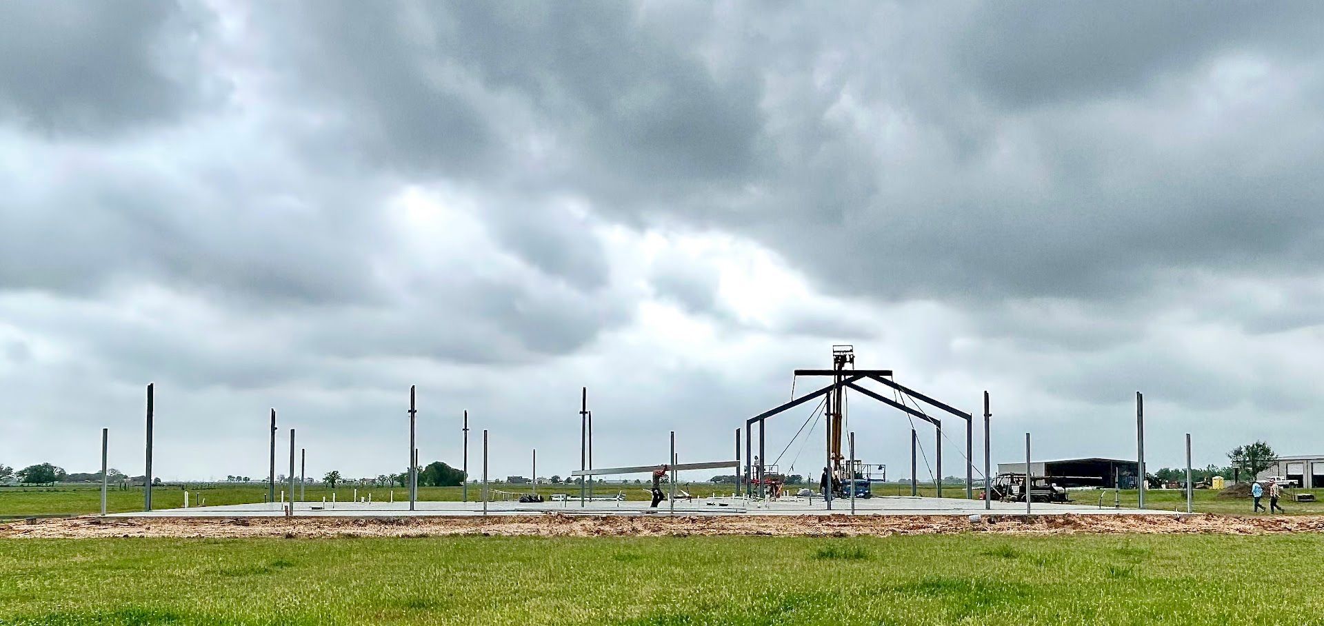 Construction site with metal framing against a cloudy sky, on a grassy field.