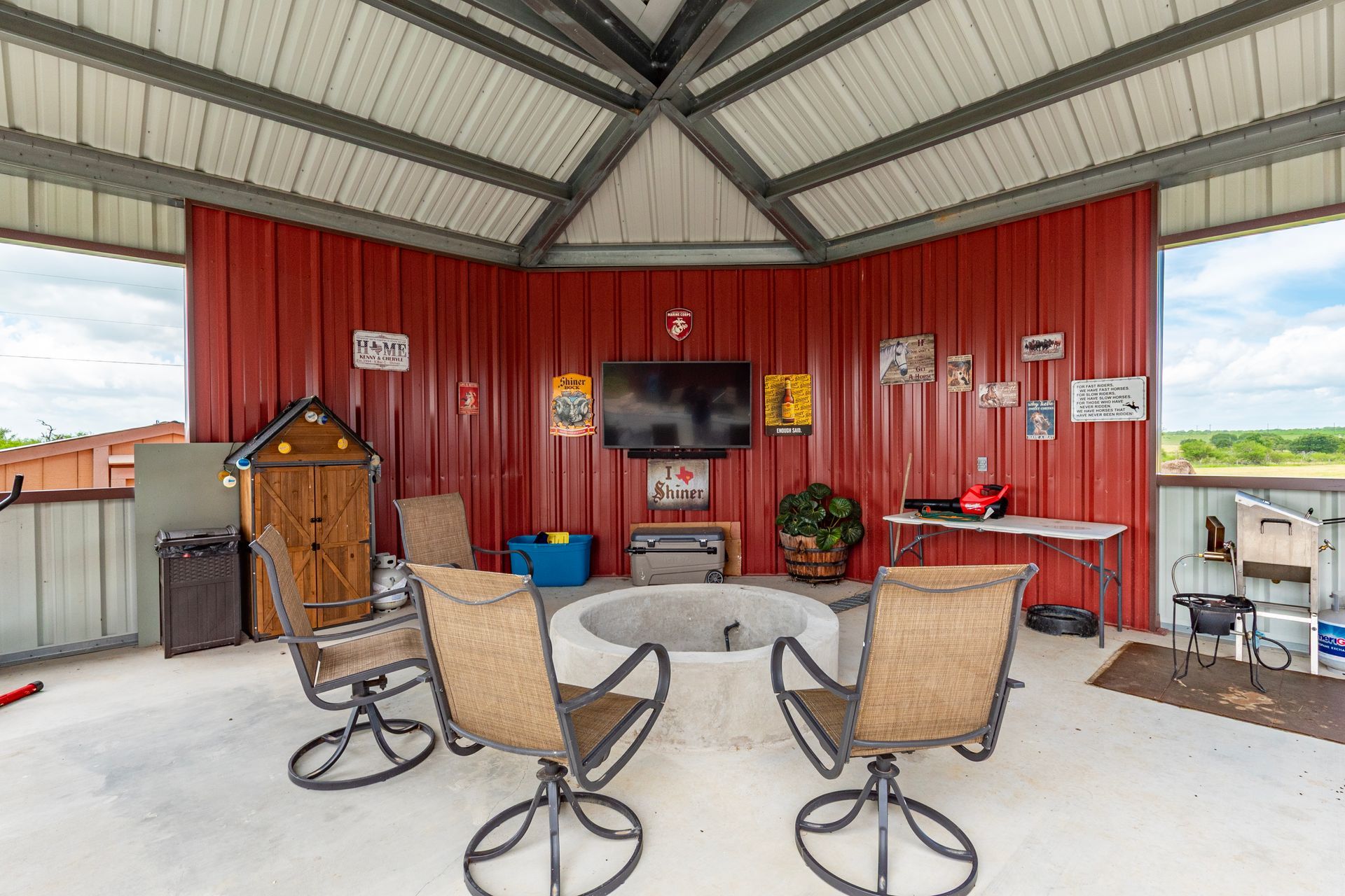Outdoor gazebo with red walls, seating around a fire pit, and a mounted TV.