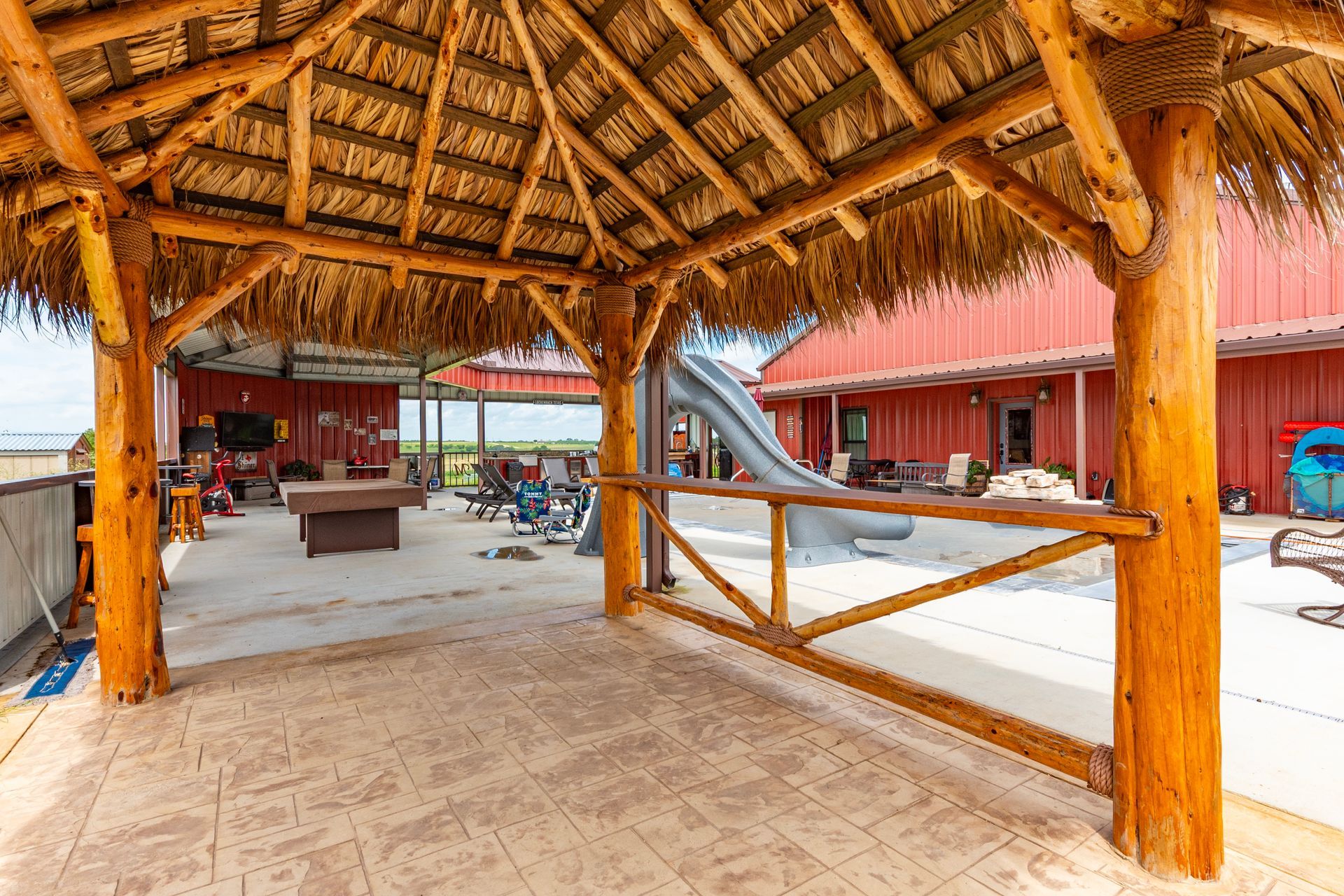 Outdoor palapa-style shelter with thatched roof, concrete patio, and red building in background.