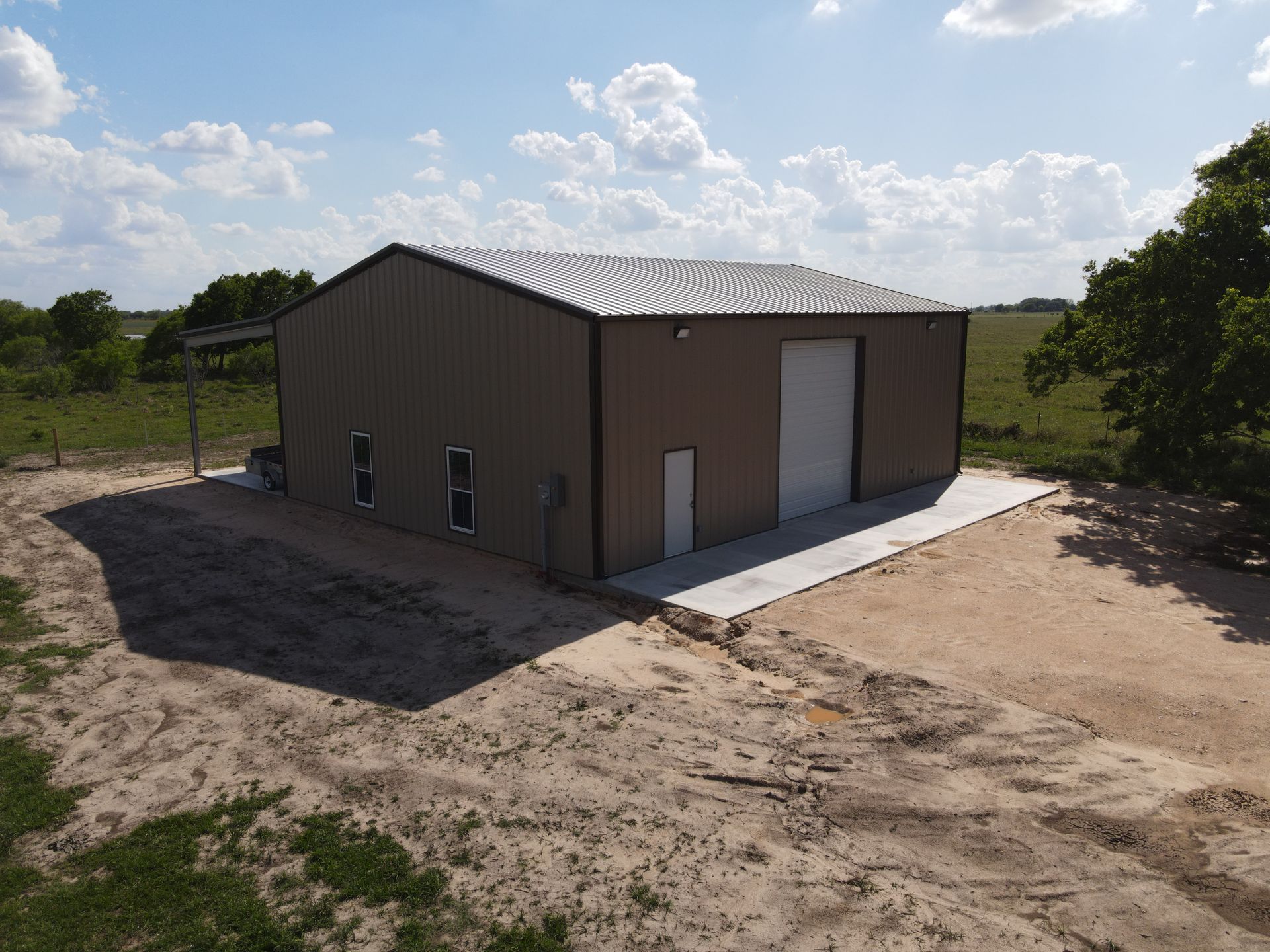 Tan metal warehouse building with concrete drive in a rural setting under a partly cloudy sky.