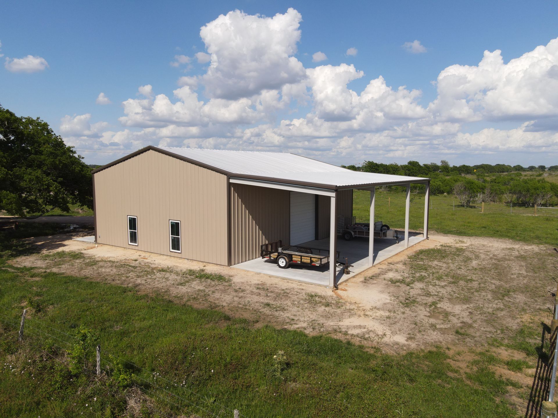 Tan metal barn with white trim and a covered patio, on a grassy rural property under a blue sky.