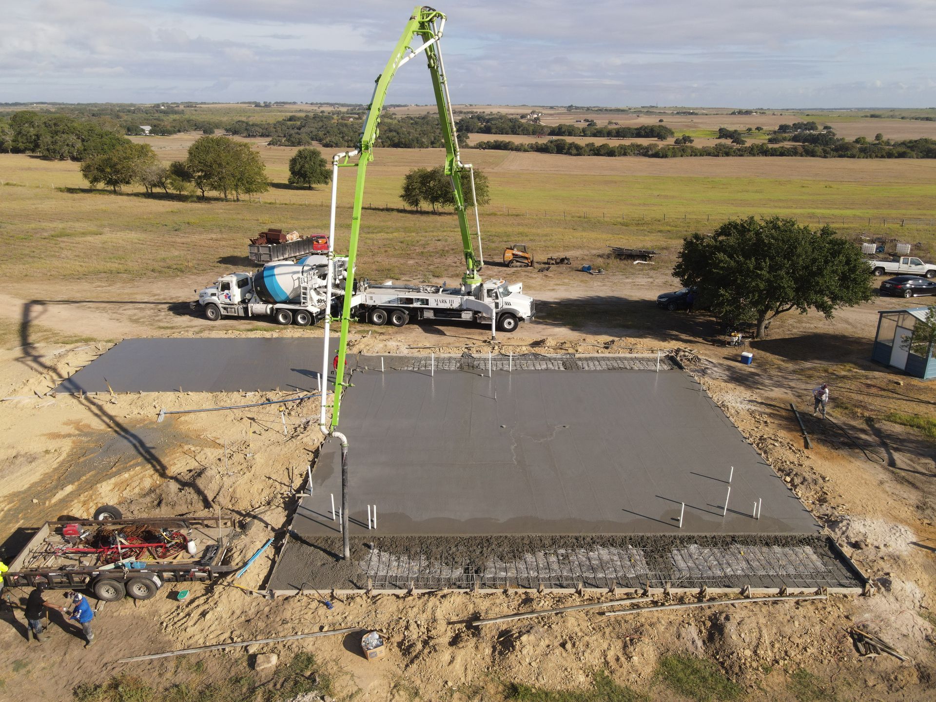 Concrete being poured on a building foundation in a rural area. A concrete pump truck and mixer are present.