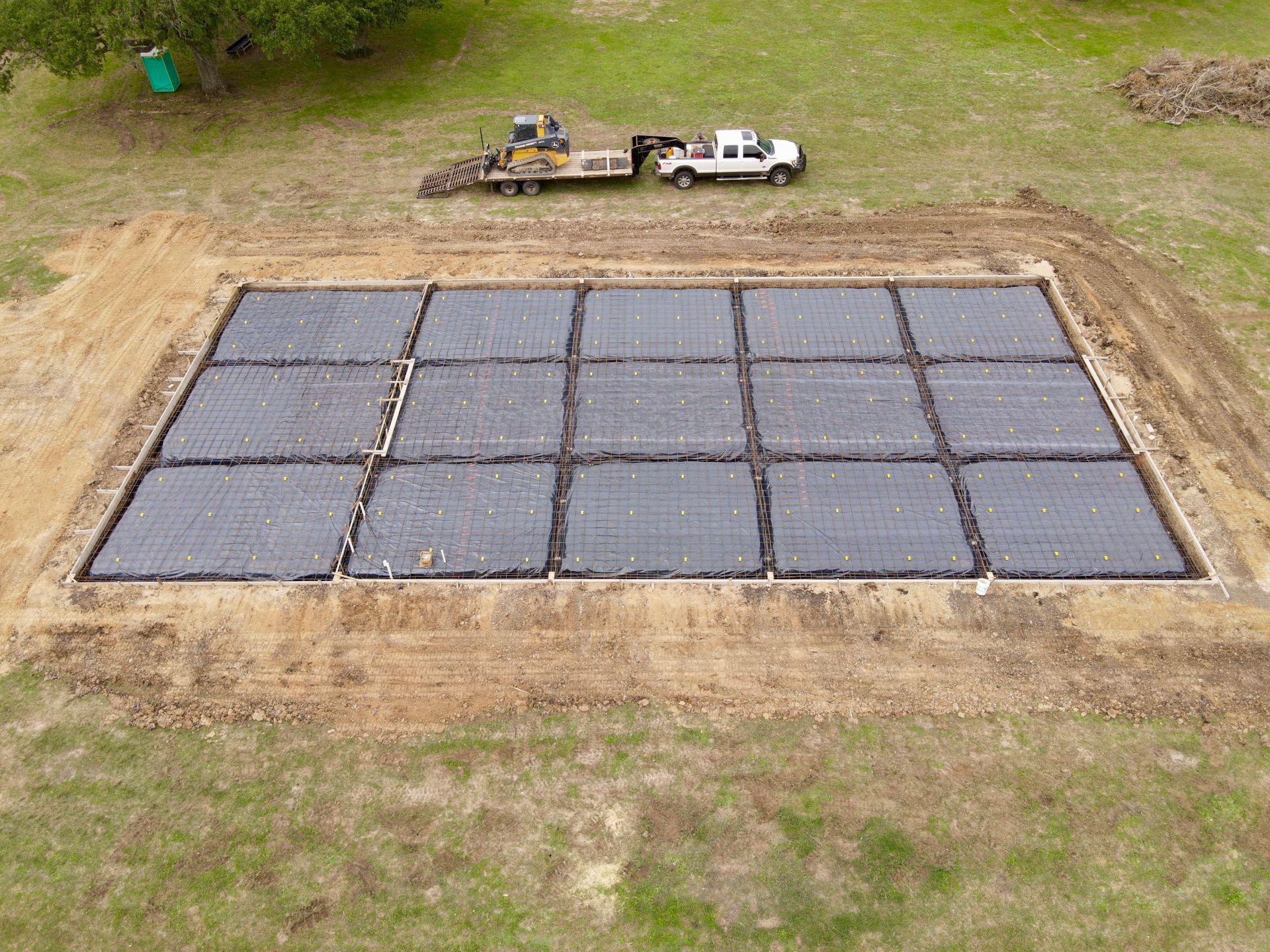Aerial view of a rectangular construction site with dark square grids, surrounded by dirt and grass, and a truck.
