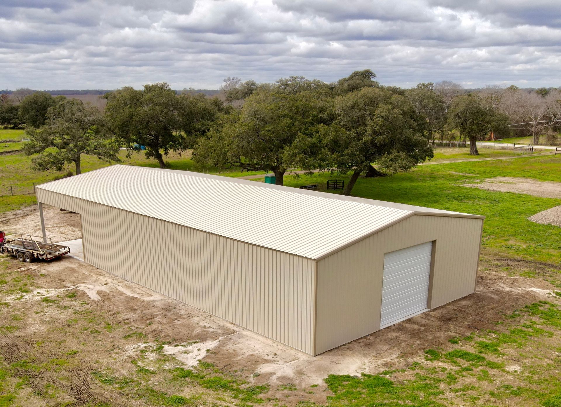 Metal barn in a field, beige with a white door. A truck sits at the side, trees in the background.