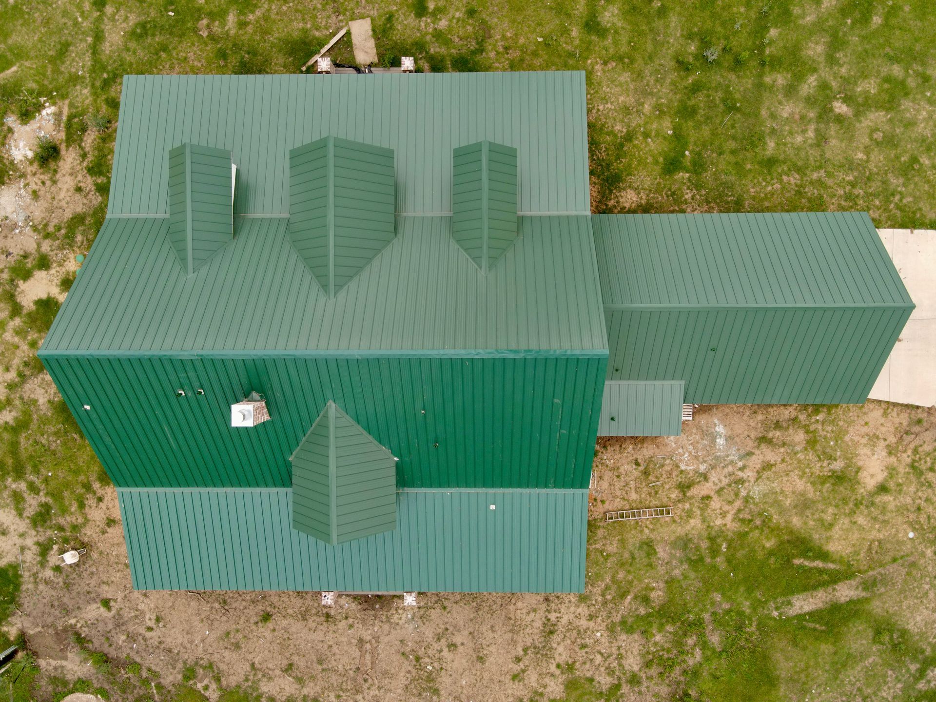 Overhead view of a green metal roof with several protruding chimney-like structures and a smaller connecting roof section on a grassy area.