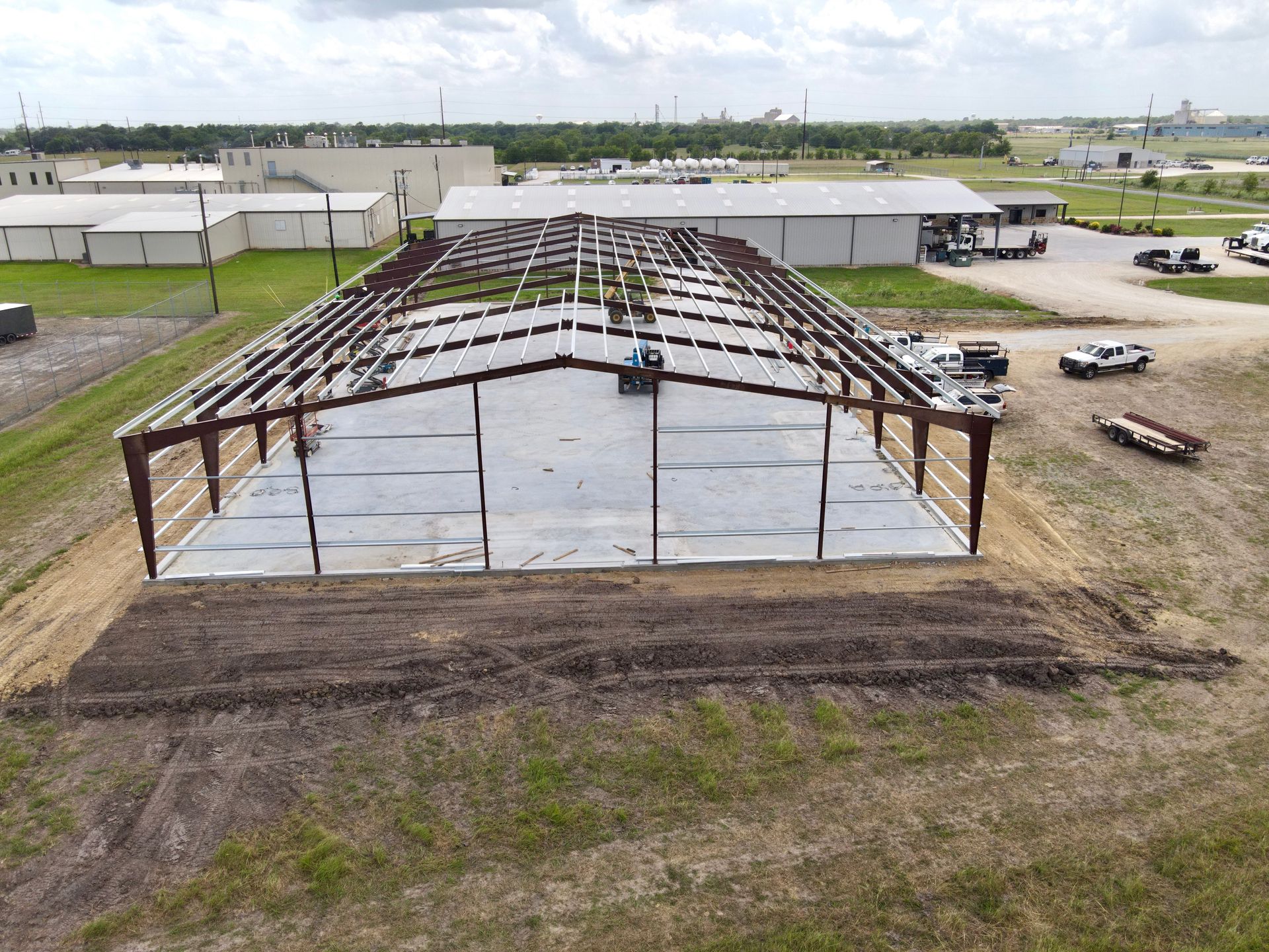 Steel frame of a building under construction on a concrete pad; rural setting with other buildings visible.