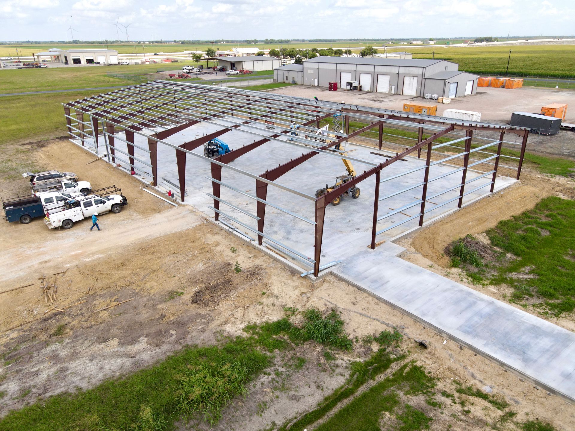 Steel frame warehouse under construction on a concrete slab, with construction vehicles and adjacent buildings in a rural setting.