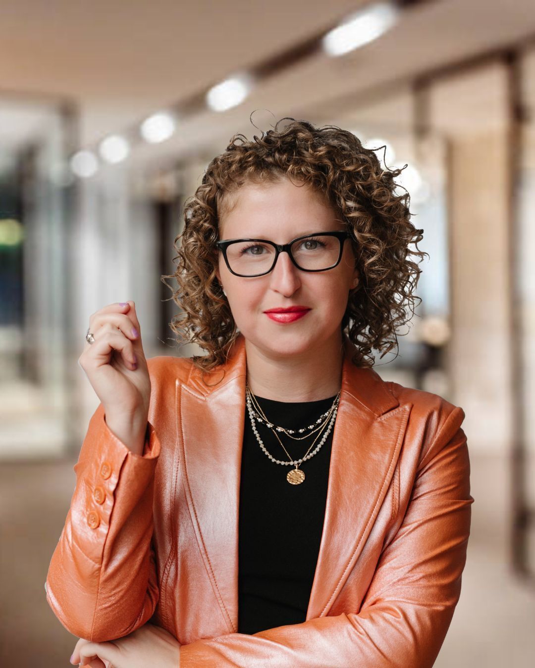 A woman with curly hair standing in a black shirt and copper jacket.