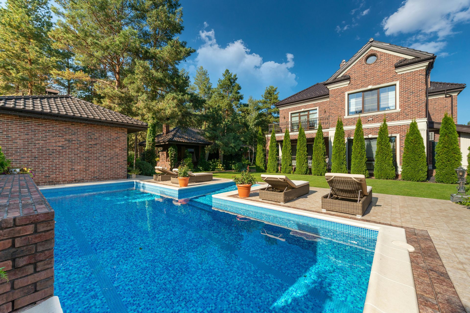 A blue pool surrounded by pool chairs, trees and a brick house