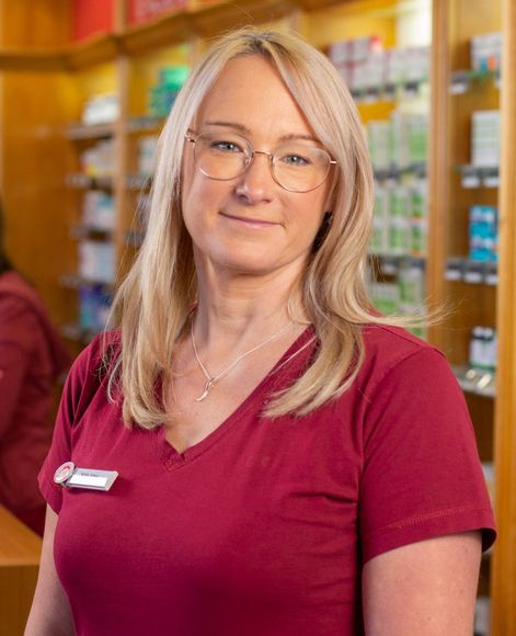 Eine Person in einem bordeauxroten Kasack mit V-Ausschnitt und Brille steht in einer Apotheke; im Hintergrund sind Regale mit Produkten zu sehen.
