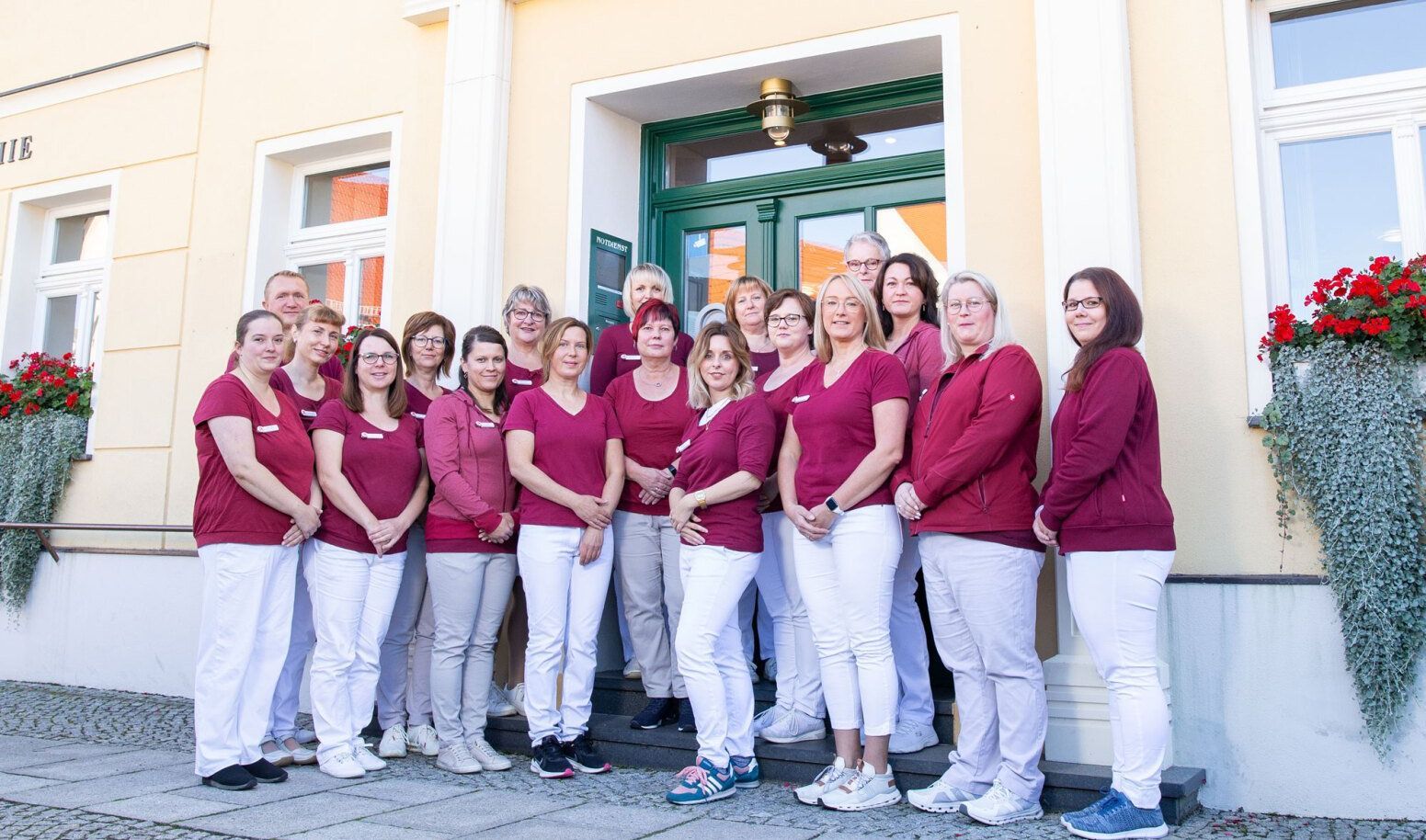 Eine Gruppe von Medizinern in einheitlichen weinroten Oberteilen und weißen Hosen steht gemeinsam vor einem Gebäudeeingang.