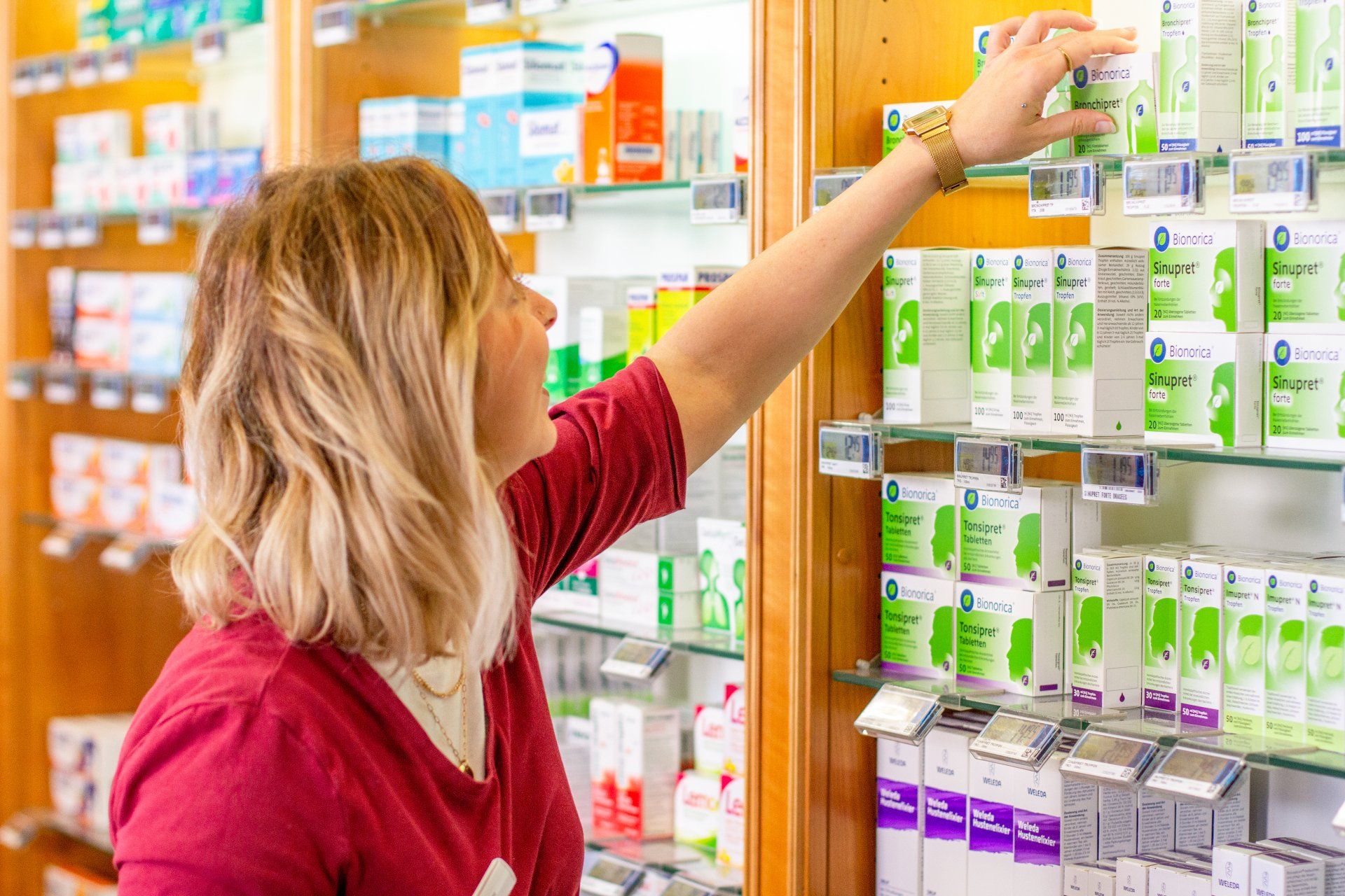 Ein Apotheker in roter Kappe greift in einer hell erleuchteten Apotheke nach einer Medikamentenpackung im Regal.