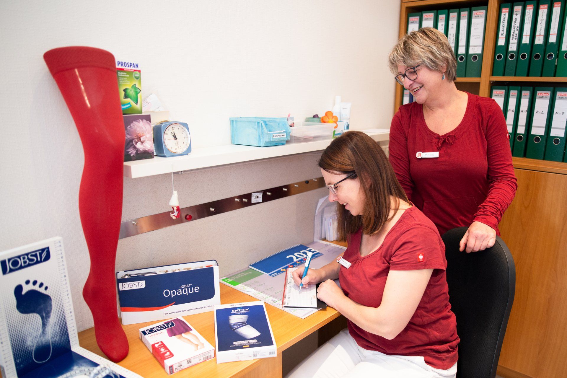 Zwei Personen in einem professionellen Büroumfeld, eine Person schreibt an einem Schreibtisch, neben dem sich medizinische Versorgungsboxen und eine rote Beinpuppe befinden.