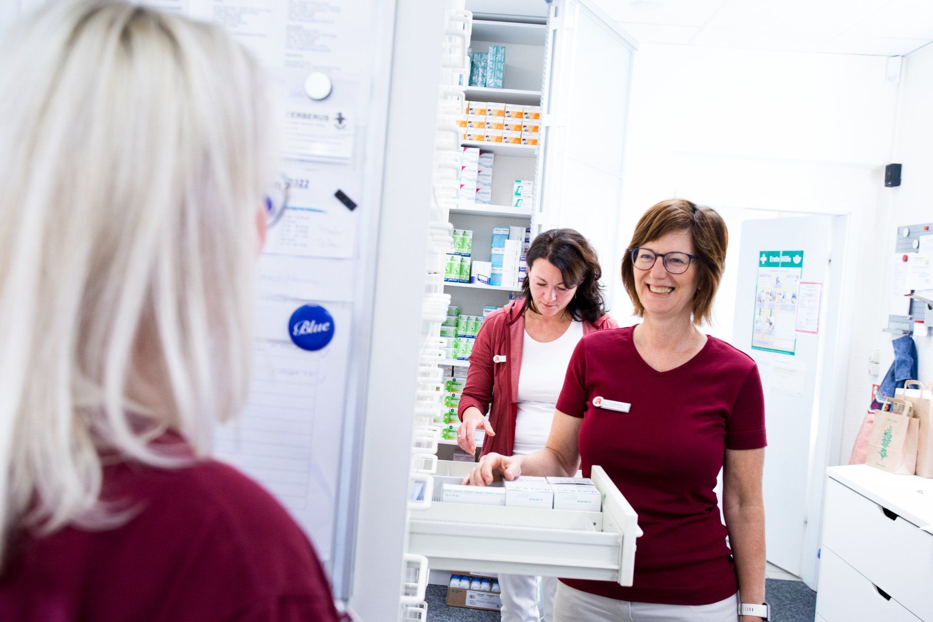 Drei Angestellte in einheitlichen bordeauxroten Uniformen arbeiten in einer hellen Apotheke mit Medikamentenregalen.