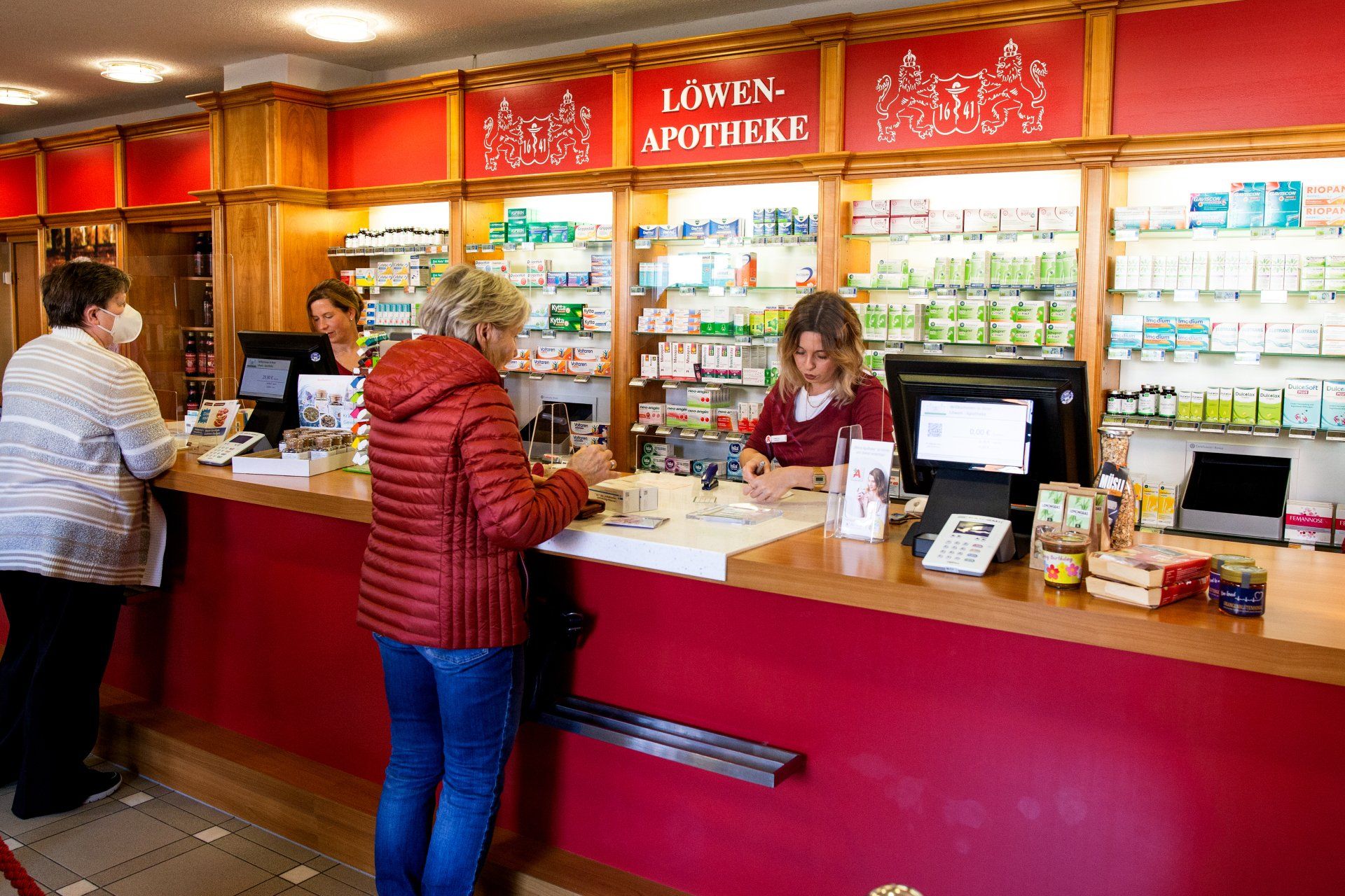 Kunden am Schalter einer Apotheke mit der Aufschrift „Löwen-Apotheke“, während hinter den Regalen Angestellte arbeiten.