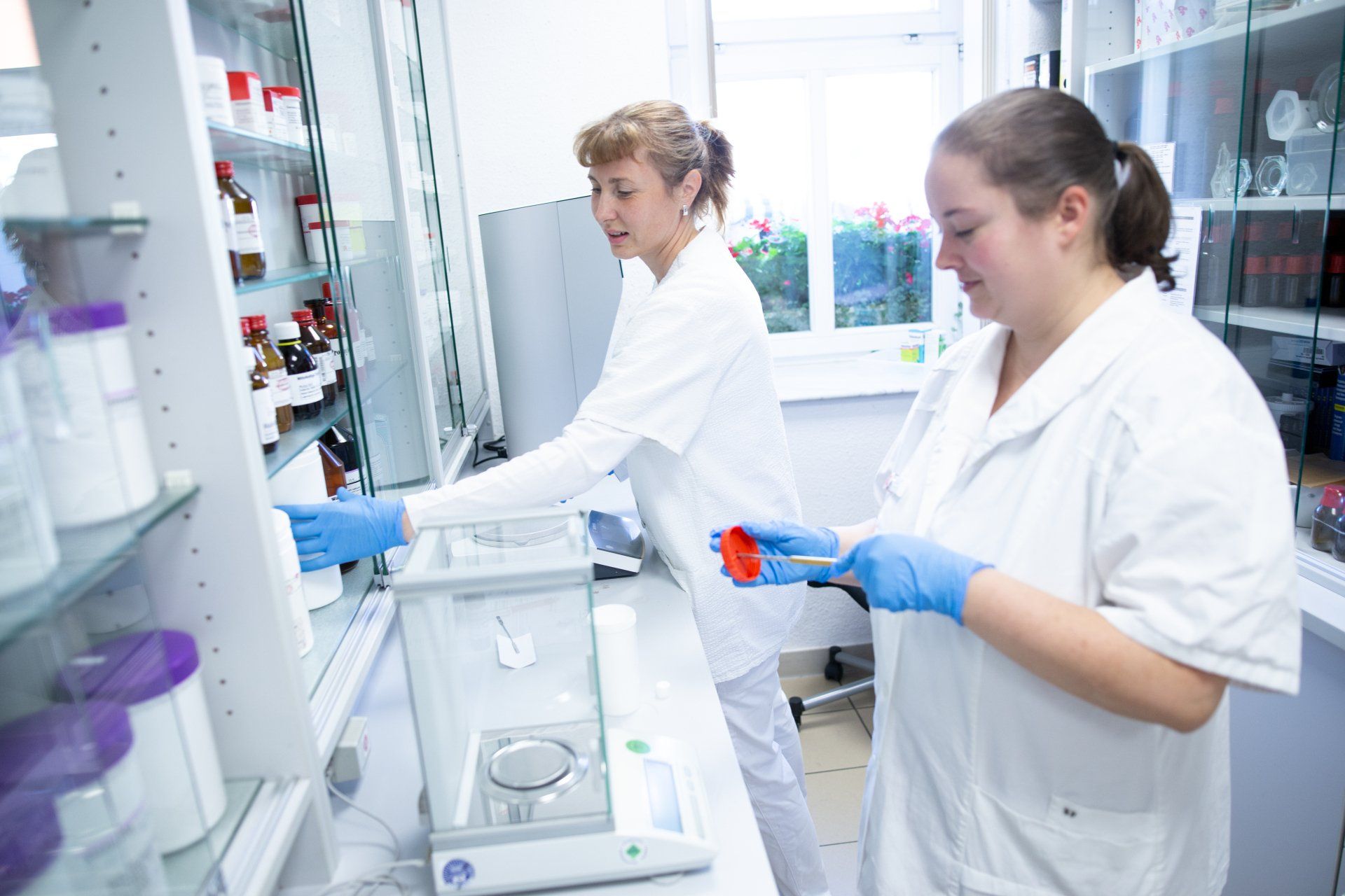 Zwei Personen in weißen Laborkitteln und blauen Handschuhen arbeiten in einem Labor und holen Geräte und Chemikalien aus einem Regal.