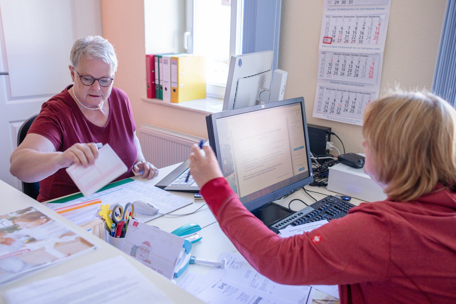Zwei Personen in roten Hemden arbeiten an einem Schreibtisch, eine hält ein Dokument in der Hand; im Hintergrund sind ein Computer und Akten zu sehen.