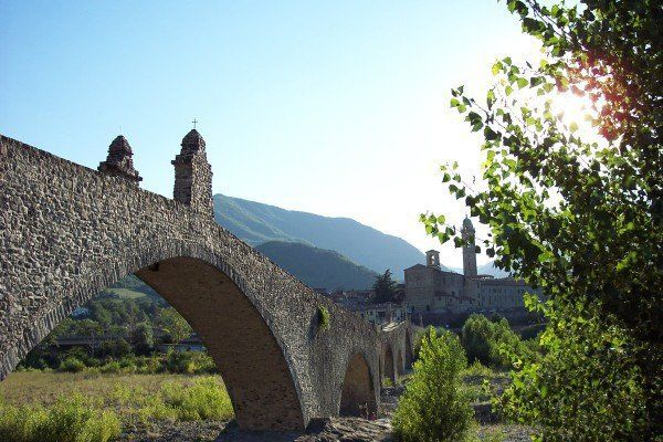 Stone arched bridge leading to a historic village with mountains in the background.