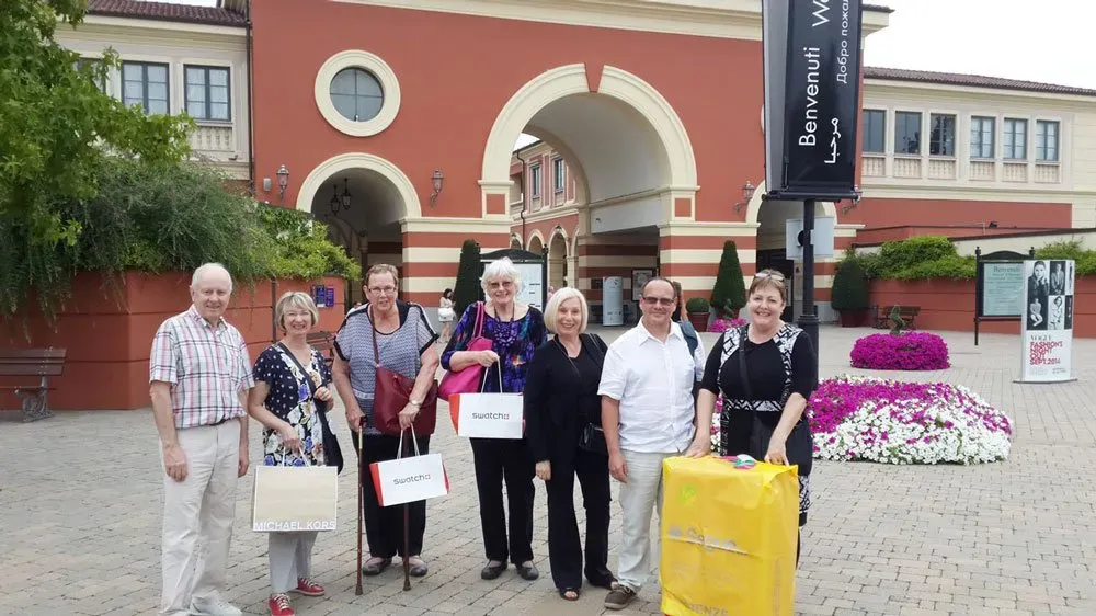 Group posing with shopping bags at a luxury outlet village entrance.