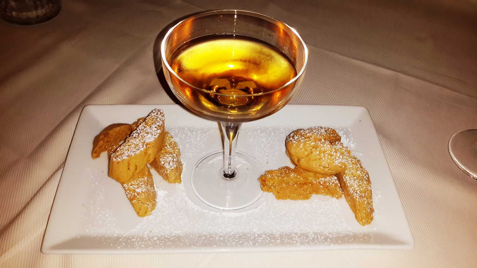 A glass of dessert wine served with biscotti and pastries on a rectangular white plate, dusted with powdered sugar.