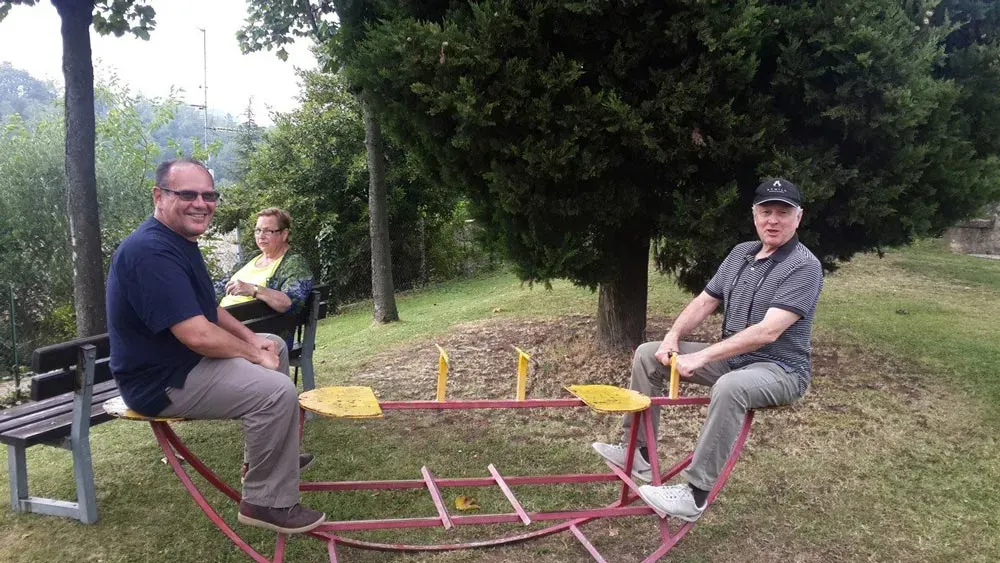 Two men playing on a seesaw in a park while a woman watches from a bench.
