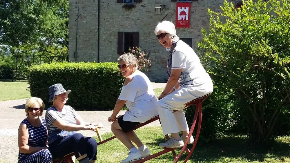 Four women playfully ride a large wooden seesaw outside a stone building under bright sunlight.