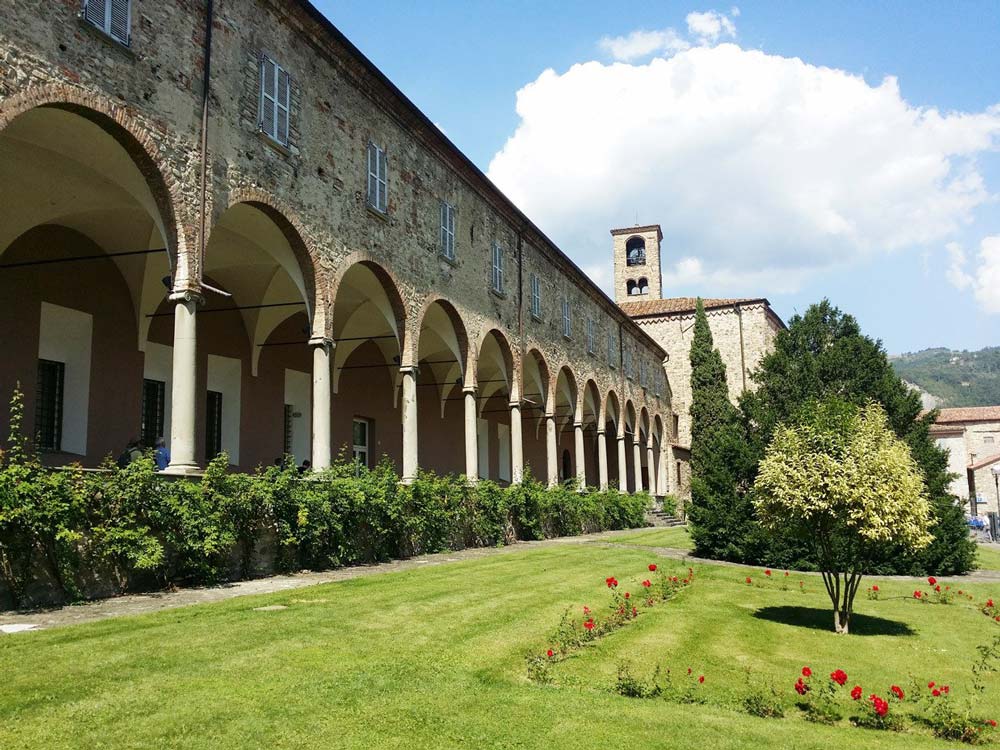 A historical stone building with arched walkways and a clock tower, surrounded by a well-maintained garden and trees.