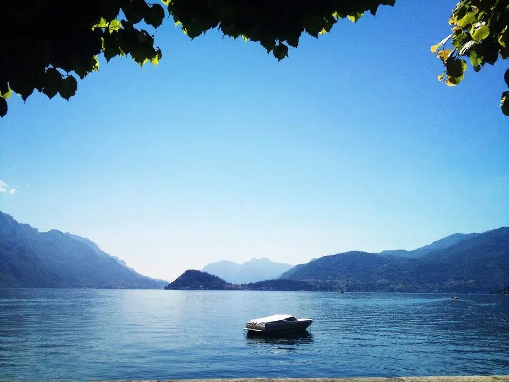 Small boat floating on a calm lake surrounded by mountains under a clear blue sky, framed by tree branches.
