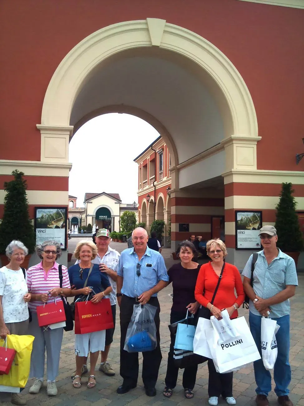 A group of people holding shopping bags standing under an archway at an outdoor shopping complex.