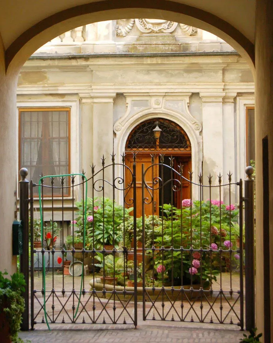 Wrought iron gate opens to a charming courtyard with lush plants and a grand wooden door framed by ornate stonework.